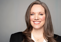 A person with long brown hair wearing a black blazer and a white top with a round pendant necklace, against a grey background.