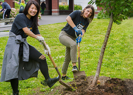 Two women dig shovels into the ground by the base of a new tree they're planting in Newark's Branch Brook Park.