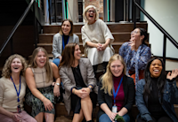 A group of Audible employees who are members and leaders of Women@Audible sit on the steps in our Innovation Cathedral building. Posed for a group photo, someone has obviously said something funny that has them all laughing and leaning into or looking at each other.