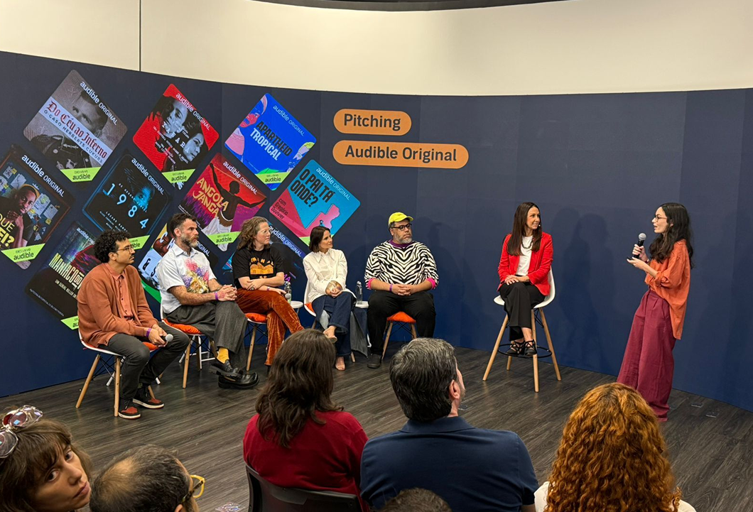 Mikaela Pasa apresenta o projeto vencedor do Pitching de sua equipe aos jurados Leo Neumann, Chico Felitti, Paula Scarpin, Bianca Comparato e MM Izidoro, sob o olhar de Adriana Alcântara, diretora-geral da Audible no Brasil.