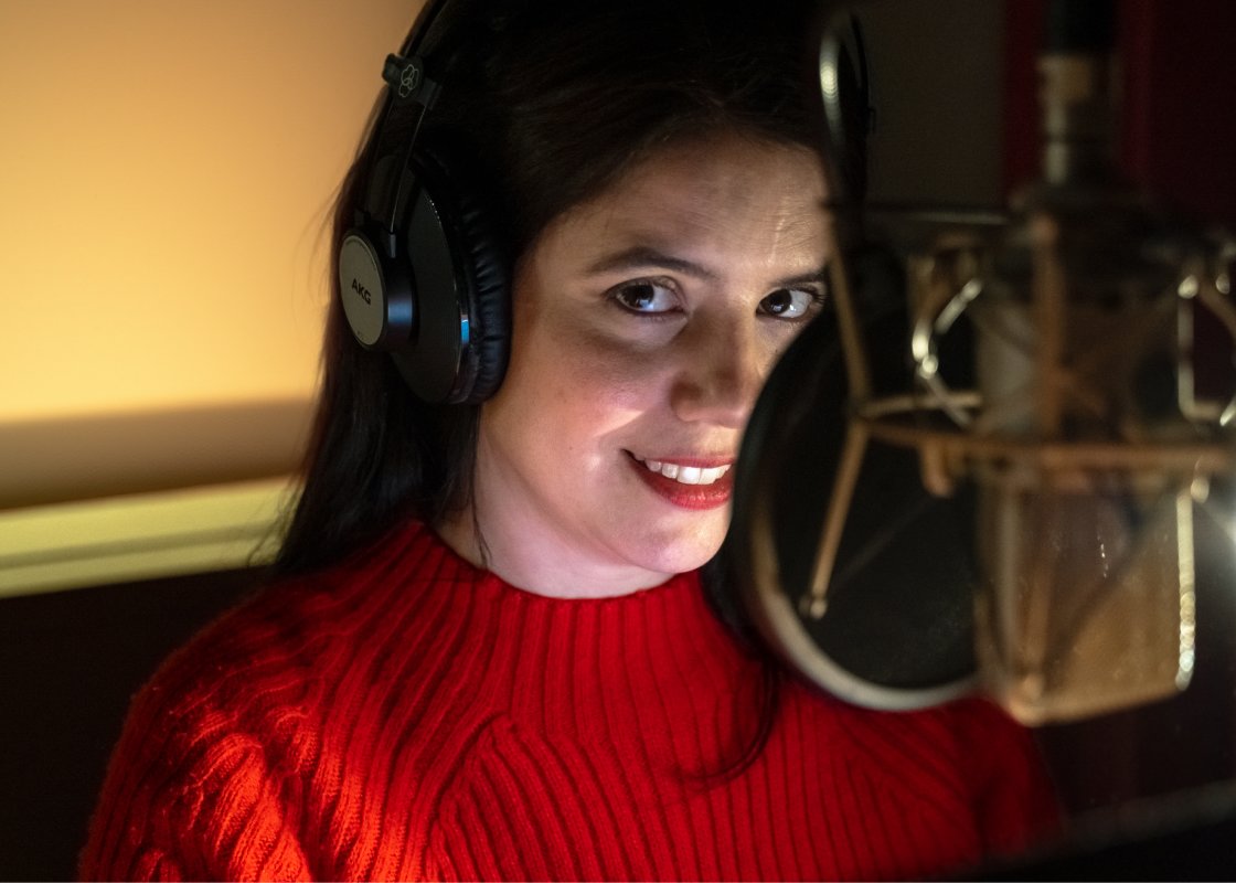 Miranda Gas stands at a microphone in a recording studio narrating an audiobook, a pair of black headphones over her ears.