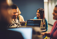 A group of Audible employees are gathered around a table in Audible's office. One woman smiles brightly to  her colleague sitting next to her.