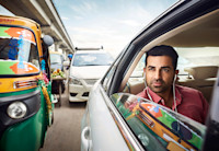 A man sits in the back of a car stuck in traffic on a road in India. His window is down and he has headphones on.
