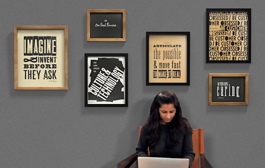 A woman working on a laptop, sitting in front of a wall of framed posters designed in a retro letterpress style, each displaying an Audible People Principle