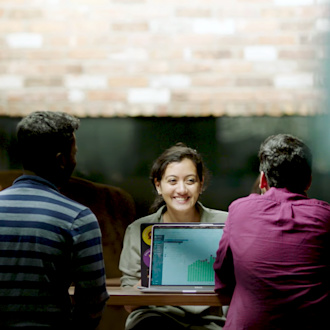 A woman sits at a conference table smiling at two colleagues who sit on the other side of the table. We see only the two men's backs. They are all working on laptops.