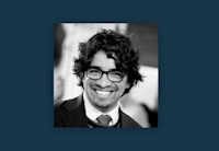 Audible Director of Spanish-Language Original Content Manny Miravete is center of image, smiling broadly. He has curly black hair and is wearing black glasses and a suit and tie. The photo is black and white and bordered by a cobalt blue frame.