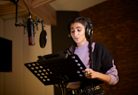 Actress Alba Flores stands in a studio recording the audio memoir "Mujer de Frontera" for Audible Spain. She has her hands holding the sides of the stand in front of her and a large microphone hangs down before her. She has headphones on and is wearing a purple and black sweater.
