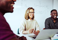 A woman sits at a conference table with two men, laptop open in front of her. All are smiling. On the white board behind her there is technical writing.
