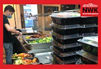Boxes of meals sit on a kitchen counter in a restaurant waiting to be delivered to Newark residents in need. Beyond them a restaurant workers is filling an empty container with chicken and greens to make up another meal for delivery.