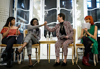 Four women sit on stage talking on a panel. The middle two women are clasping hands in a type of high five.