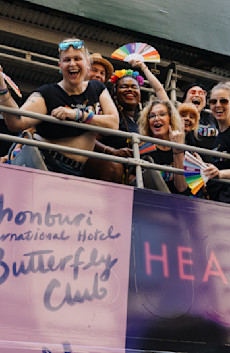 Audible creators and talent cheer and hold rainbow-colored fans on top of the Audible-branded double decker bus. Artwork for the Audible Original "Chonburi International Hotel Butterfly Club" is seen below them.