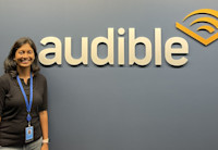 Audible employee Nandhini Gunalan, who participated in the 'New Chapter' Returnship Program, stands in our Newark office by an Audible logo on the wall. She is wearing a black shirt and her ID and lanyard and smiling.