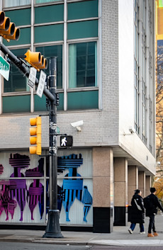 a city street corner with persony colorful striped figures on storefront windows, street lights and people walking
