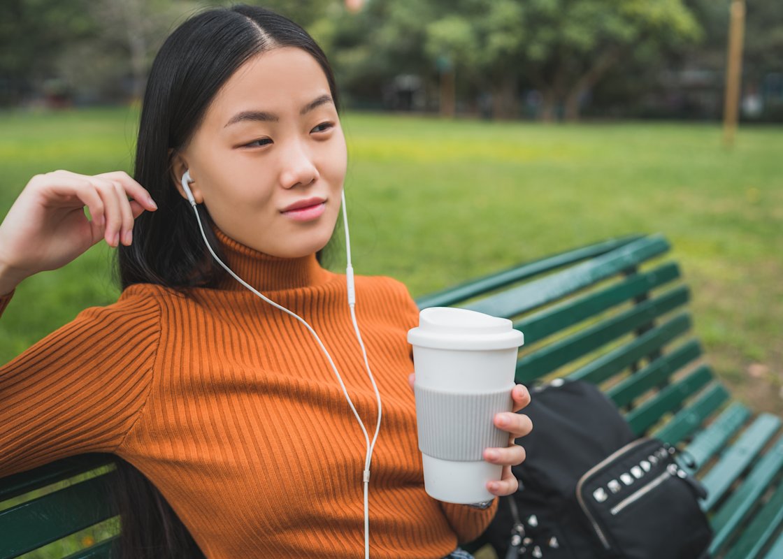 A woman sits on a park bench listening to an audiobook through the headphones in her eats while holding a coffee cup. Her bag sits on the bench beside her.