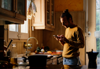 A photo of a man standing by a kitchen sink. He's looking at his smartphone, selecting an option on it.