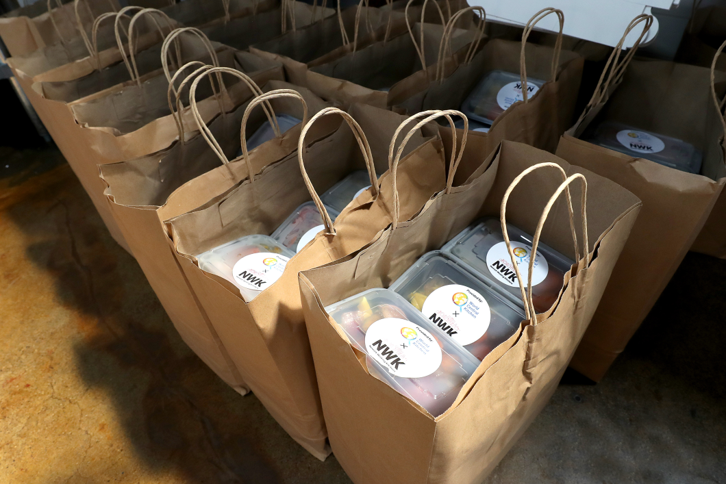 Brown paper bags with handles sit set up in a row and filled full of meals packaged in plastic containers with NWK logos on them. 