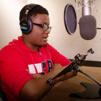 One of Audible's high school interns sits in a recording studio in front of a large hanging microphone. He is wearing glasses and a red sweatshirt and reading aloud from a text in front of him to test out what it's like to be an audiobook narrator.