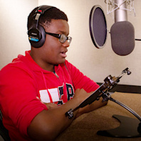 One of Audible's high school interns sits in a recording studio in front of a large hanging microphone. He is wearing glasses and a red sweatshirt and reading aloud from a text in front of him to test out what it's like to be an audiobook narrator.