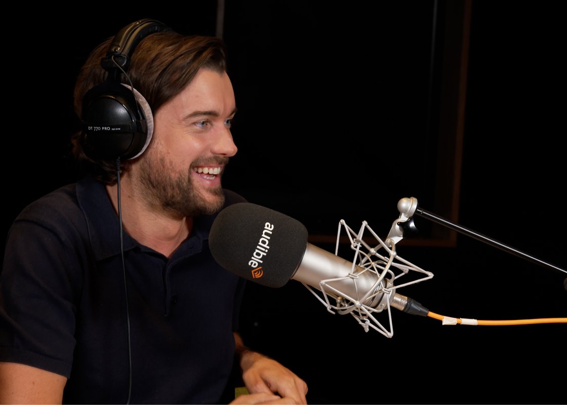 Jack Whitehall sits in front of a microphone in an Audible recording studio, black headphones over his ears.