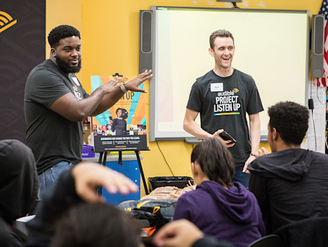 Two Audible employees stand at the front of a classroom presenting Project Listen Up to high school students seated at desks.