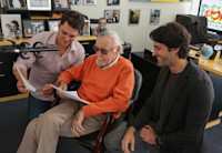 Stan Lee sits in front of a microphone reviewing text with two collaborators