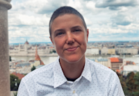 Audible employee Lisa Del Monte sits on the edge of a stone balustrade looking direct to camera while wearing a black and white polka-dotted buttoned down collared shirt. Behind Del Monte, a city can be seen with a wide river flowing through it and white buildings with orange domes.