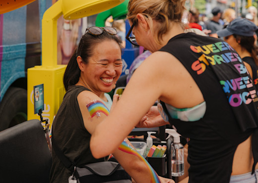 A woman smiles in joy while getting a rainbow painting on her arm. The artist has her back facing the camera, and you can read "Queer Stories by Queer Voices" on the back of her shirt.