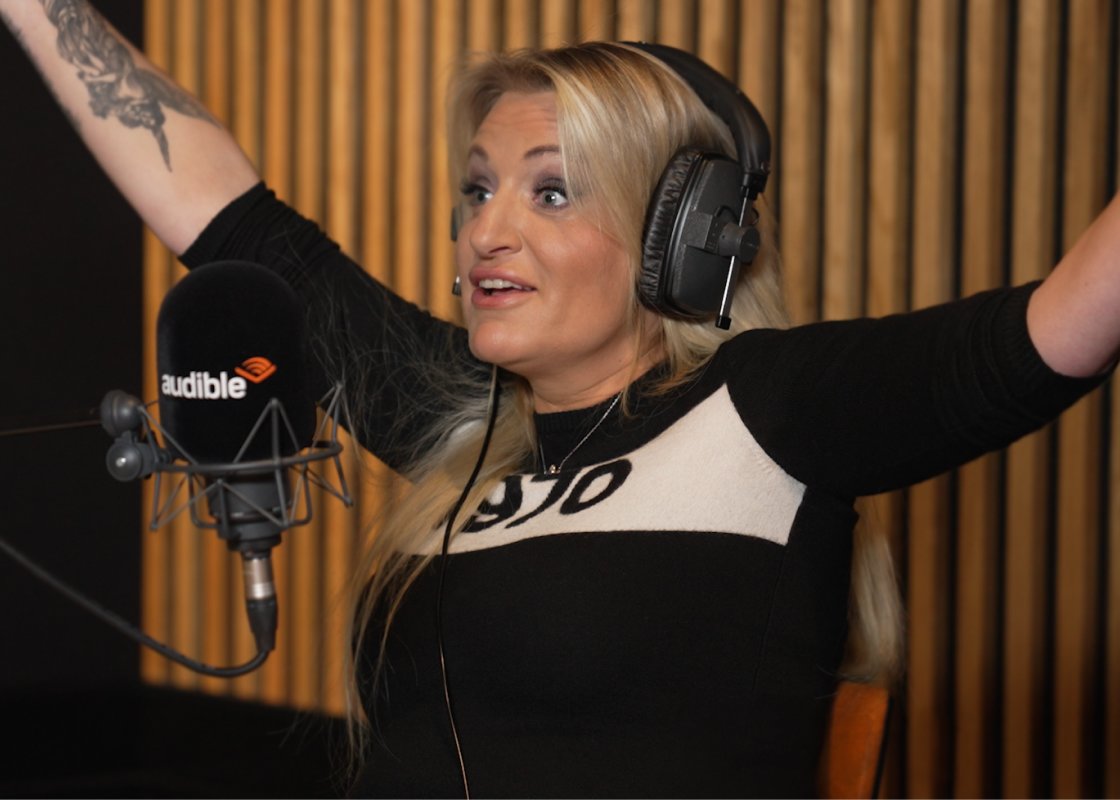 Daisy May Cooper stands at the microphone in an Audible studio, recording her podcast, her arms outstretched over her head.