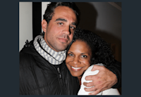 Actor Bobby Cannavale stands with his arm around actor Audra McDonald, holding her close. She is smiling and they are both looking directly into the camera. He is wearing a black zip up jacket with a gray and white scarf wrapped around his neck. She has on a white long sleeve shirt.