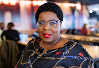 Audible employee Marlesha Ross stands in the front of the frame wearing a blue shirt with gold links designed on it with Audible's large cafeteria in its Newark office blurred in the background.