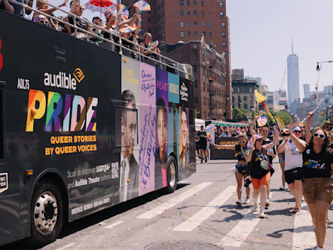 The Audible branded double decker bus travels down the streets of New York City. Audible community members walk in the parade waving flags to celebrate Pride Month,