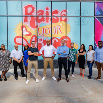 Group of people wearing business casual attire smile in a straight line. Behind them is a colorful window façade that says "Raise Your Voice."