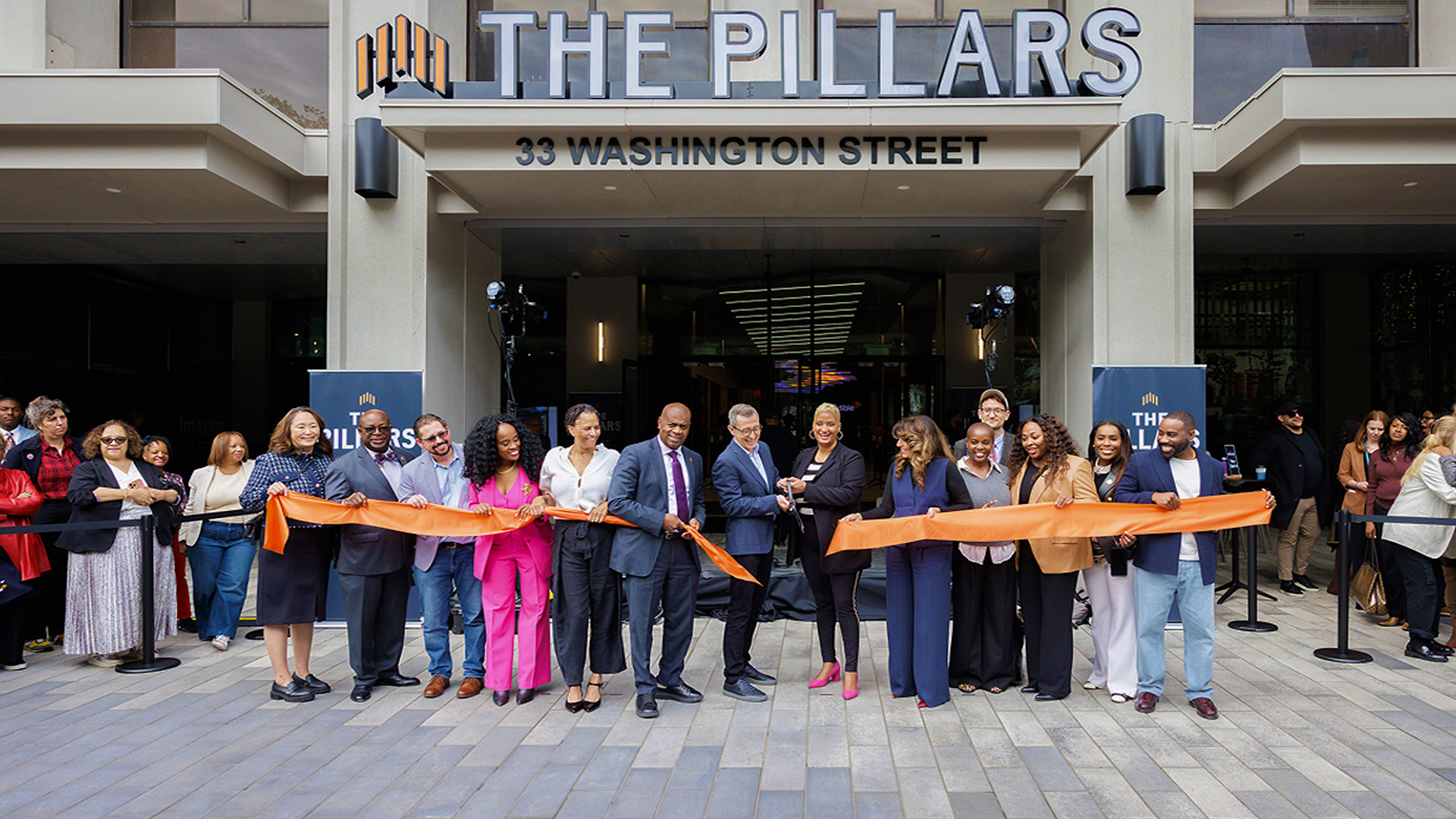 A crowd is gathered in front of a building called "The Pillars" in downtown Newark with people cutting a large orange ribbon to mark its opening.