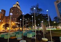 Solar lights and green hexagon planters and seats are artfully arranged in Washington Park with Audible's headquarter buildings lit up in the background.