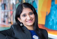Audible employee Akanksha Gupta sits in Audible's Newark headquarters lobby wearing a blue shirt and grey blazer. She is looking straight at the camera and is smiling. Behind her is a multi-colored mural that is blurred to keep her the main central focus.
