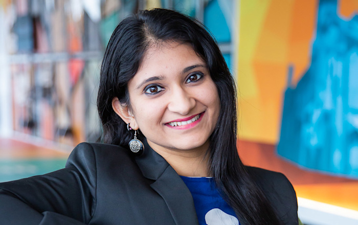 Audible employee Akanksha Gupta sits in Audible's Newark headquarters lobby wearing a blue shirt and grey blazer. She is looking straight at the camera and is smiling. Behind her is a multi-colored mural that is blurred to keep her the main central focus.