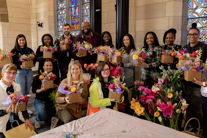 A large group of Audible employees are posing in a group with floral arrangements from a team building event. They are standing in front of a stained glass window in the Innovation Cathedral.