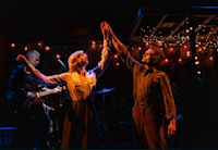 A photo of a scene from "Dead Outlaw." Performers Julia Knitel and Andrew Durand are illuminated while dancing in dimly-lit room. A guitarist is visible in the background.