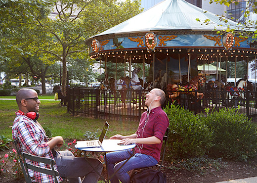 Two men work on computers at a table in a park in Newark with their headphones on. A carousel is behind them.