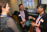 Three women stand talking in a group. They're smiling and animated. In the background is a bit of a staircase and stained glass wall from Audible's Innovation Cathedral, a restored church that now operates as a office.