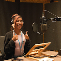 A student sits in a recording studio, headphones on, mic in front of her, making the thumbs up sign.