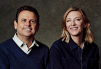 Cate Blanchett and Danny Kennedy sit side-by-side against a grey background. They are looking at the camera and smiling. Both are wearing dark blue, Blanchett in a collared blue button down and Kennedy in a blue v-neck sweater over a cream button down shirt with small fish patterned on it.