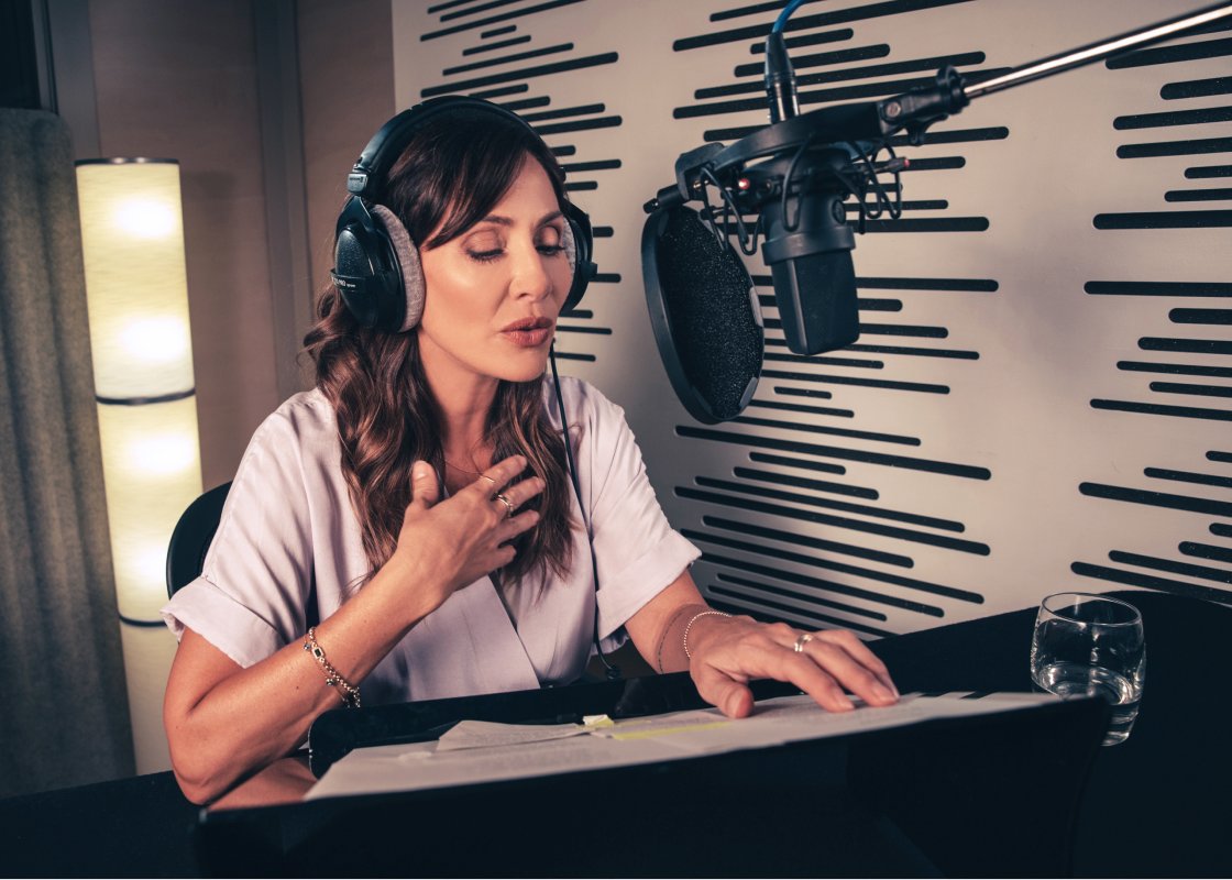 Natalie Imbruglia Sits in a recording studio in front of a microphone, a script open in front of her, and headphones on her head.