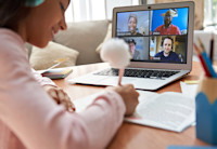 A girl sits at a table in her house in front of a laptop with her headphones on. She is making notes on a book she's reading along with four Audible employees shown on her laptop screen.