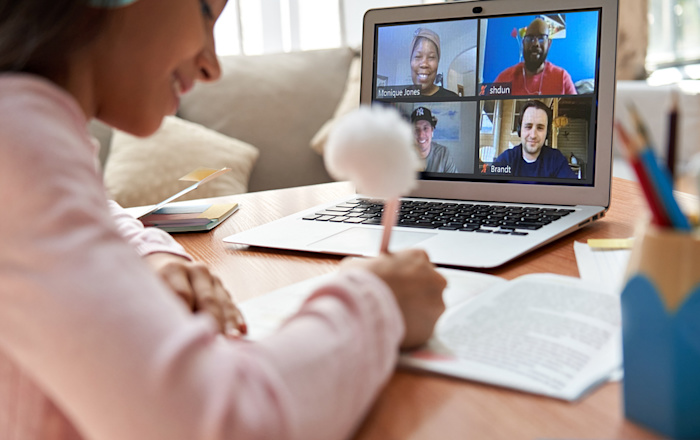 A girl sits at a table in her house in front of a laptop with her headphones on. She is making notes on a book she's reading along with four Audible employees shown on her laptop screen.