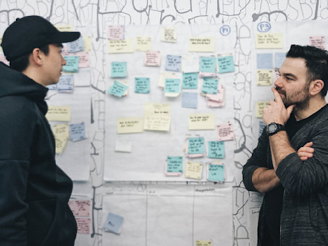 Two men stand facing each other and talking in front of a board covered with different colored post-it notes, all with writing on them. The man on the left is wearing a gray hoodie and black baseball cap and the man on the right is wearing a black shirt and gray zip on sweater.