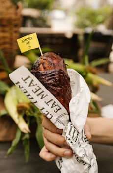 A person's hand holding a turkey leg wrapped in a wrapper that says "Eat Me" and with a flag on it saying "Smoky AF"