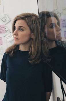 A woman with long blonde hair and with a black crew neck shirt on leans against a wall, her face reflected in the screen next to her, looking off in the distance with a wall of post-it notes behind her.