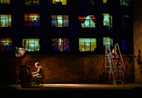 On a theatre stage, Jake Gyllenhaal sits playing a piano downstage right with a large light on him while Tom Sturridge sits on a rolling ladder upstage left looking out at the audience. An image is projected on the wall behind them to make it look like an apartment building with lots of lit windows framed by curtains.