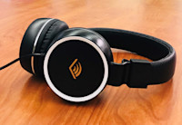 A pair of headphones rest on a wooden table top. The headphone are black and the over-the-head variety. On the outside of the cushioned earphones there is the orange chevron from the Audible logo.